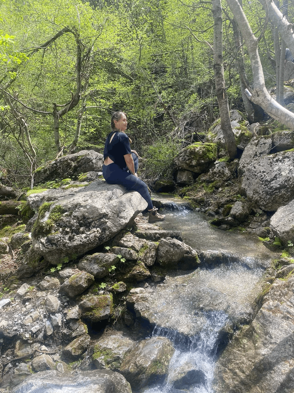 Zu sehen ist ein Bachlauf der sich durch kleine Felsen schlängelt. Das Wasser fließt ruhig und spiegelt das Sonnenlicht in sich. Eine Frau sitz auf einem großeren Fels und genießt die Ruhe der Natur. Man sieht sie von der Seite. Ihre Harre sind in einem geflochtenen Zopf zusammengefasst und sie trägt eine dunkle Leggings und ein dunkles Oberteil. Im Hintergrund stehen viele Bäume und bilden ein dichtes Blätterdach