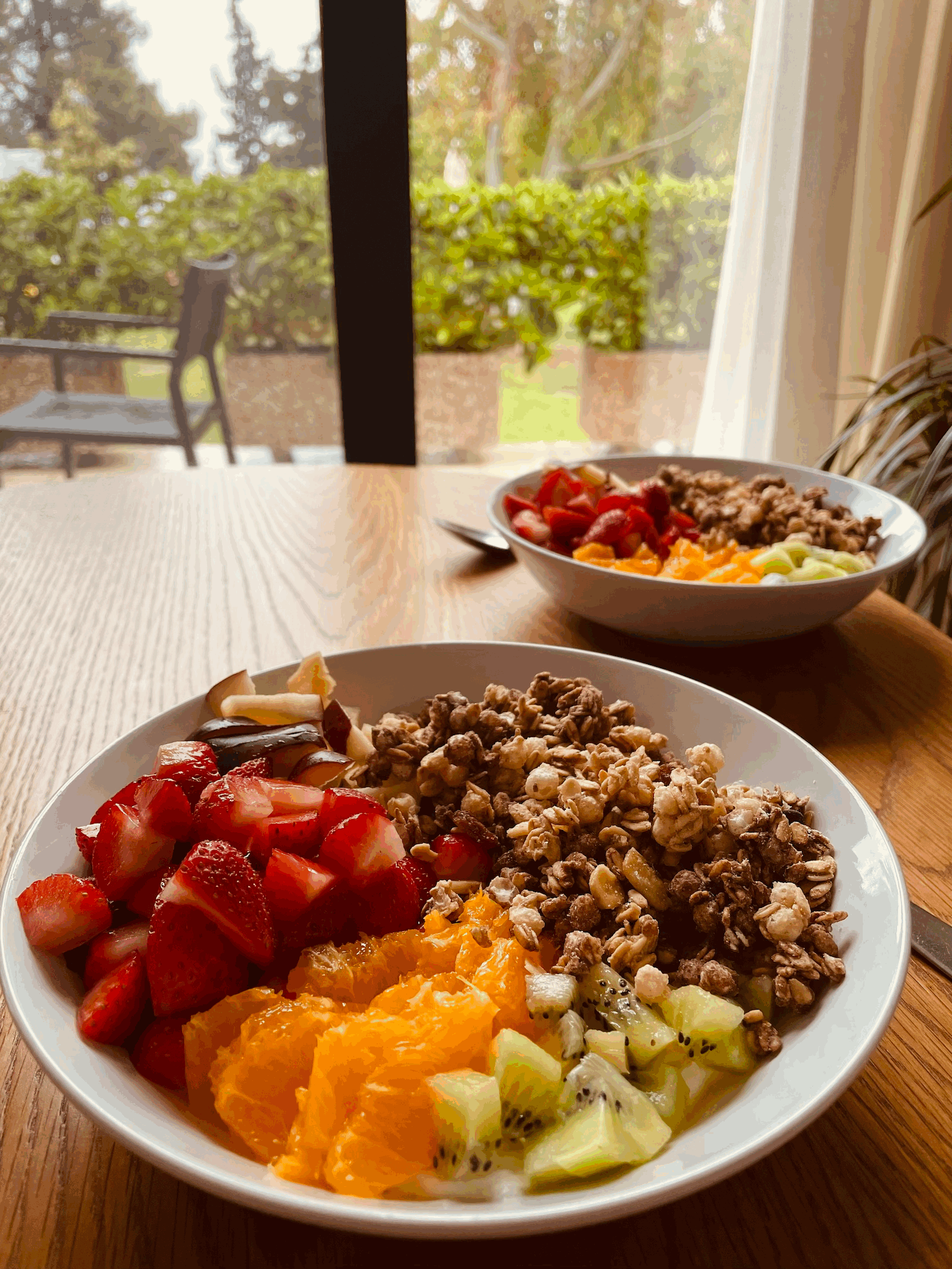 Auf dem Foto sind zwei Frühstücksschalen zu sehen. Beide haben denselben Inhalt. Das Obst in einem Bogen angerichtet in der folgenden Reihenfolge: Kiwi, Orange, Erdbeere, Apfel. Es sieht aus wie ein Regenbogen! Daneben liegt auf dem Joghurt ein Crumble Müsli.