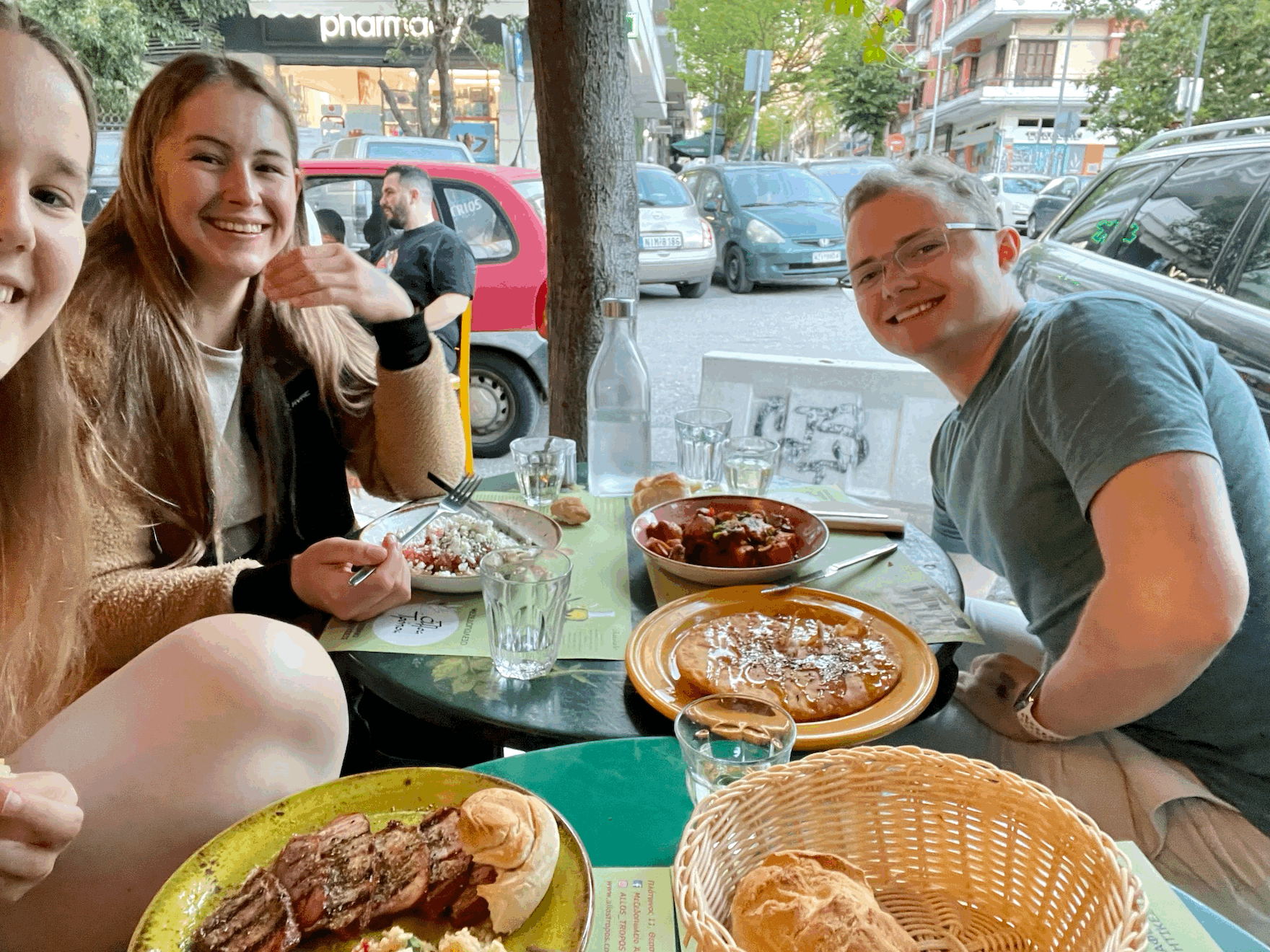 Auf dem Selfie ist das Gesicht der Fotografin abgeschnitten, sodass man nur noch ihr blondes Haar sehen kann. Sie sitzt mit zwei weiteren Personen an einem Esstisch. Vor ihr steht ein Teller mit Essen, der ebenfalls kaum zu sehen ist. Direkt hinter der blonden Frau ist eine Frau mit braunem Haar zu sehen. Sie lächelt in die Kamera und hält eine Gabel in der rechten Hand. Vor ihr steht eine Schale eines griechischen Salats. Ihr gegenüber sitz ein junger Mann in blauen t-Shirt. Auch er lächelt in die Kamera. Vor ihm stehen auch zwei Gerichte. Eines ist mit Fleisch und Gemüse, das andere ist eine griechische Nachspeise: Ein Fladen mit Honig beträufelt und Feta gefüllt.