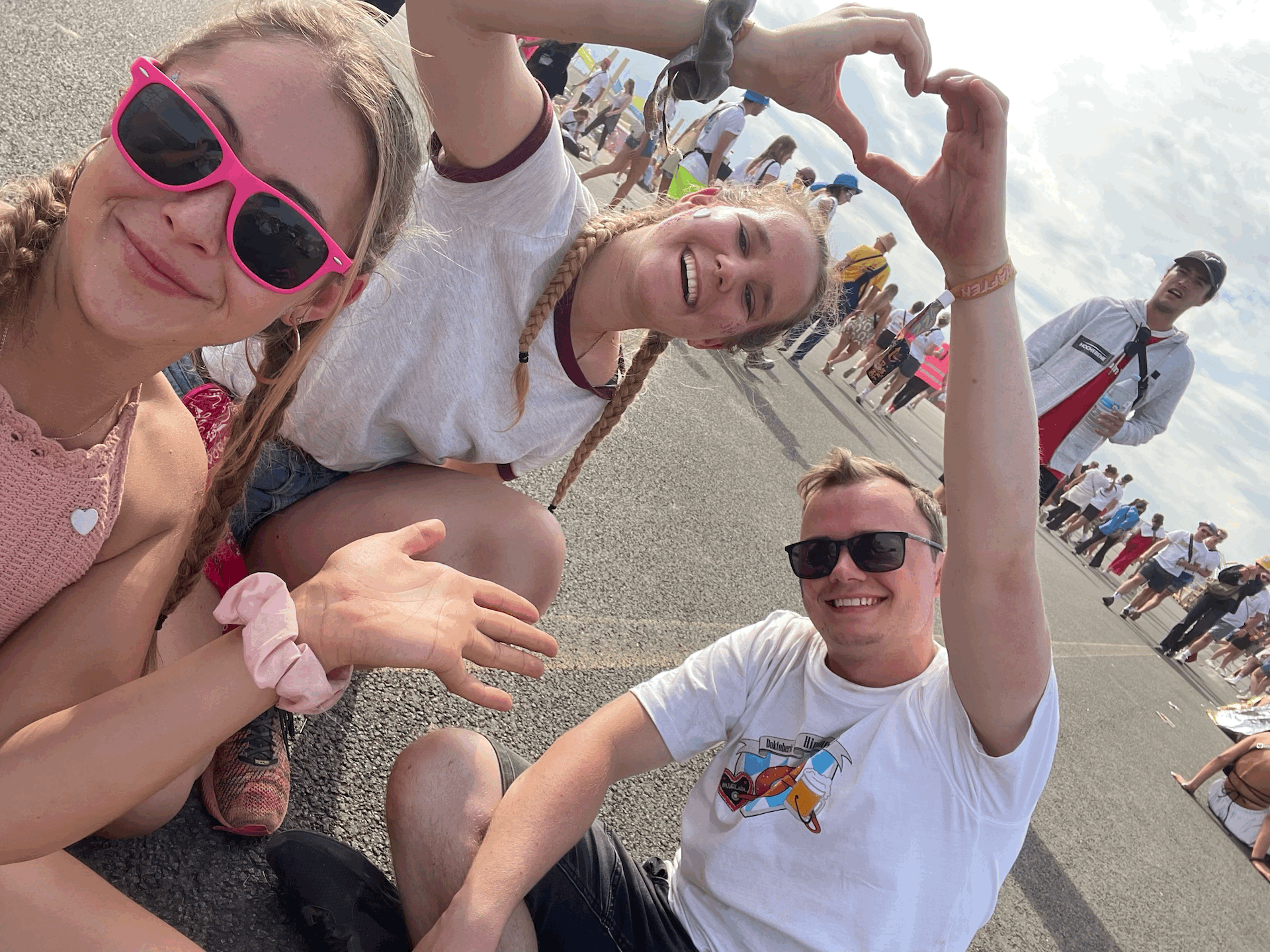 Die Fotografin trägt eine rosa Brille und ein rosé farbenes Shirt. Mit ihrer Hand deutet sie auf die zwei Personen hinter ihr, die mit ihren Händen ein Herz über ihren Köpfen formen. Alle sitzen sie auf einer asphaltierten Fläche und lächeln in die Kamera. Die zwei hinteren Personen, ein Junge und ein Mädchen, tragen beide helle Oberteile. Im Hintergrund sind viele Menschen zu sehen.