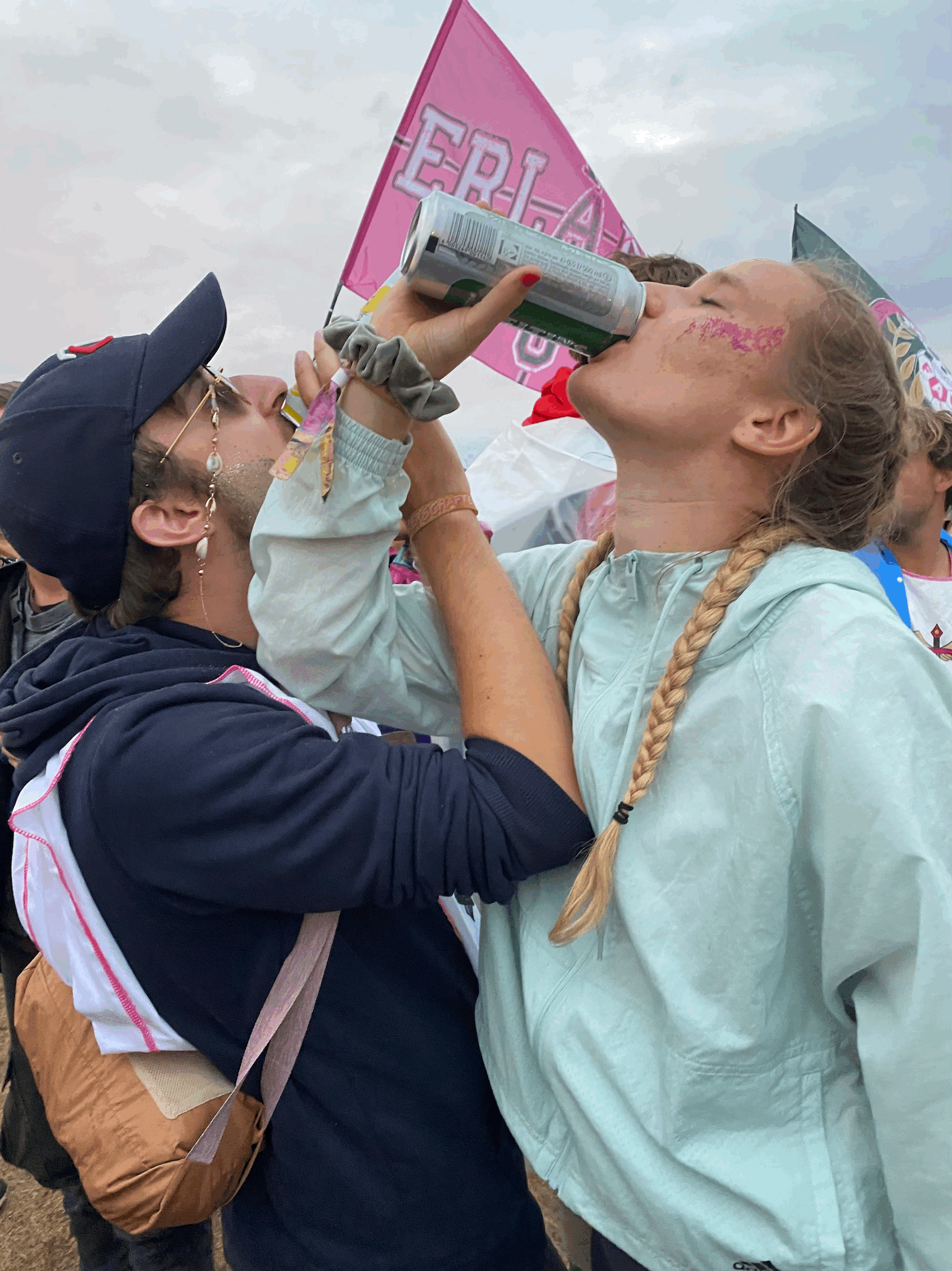 Auf dem Bild sieht man zwei Personen im Vordergrund. Ihre Arme sind verschränkt, sodass sie mit ihrer Hand ein Dosenbier trinken können. Rechts steht eine Frau in einem hellblauen Windbreaker, ihre Haare sind in zwei Zöpfen geflochten und auf ihrer Wange glitzert rosa Glitter. Die zweite Person ist ein Mann. Er trägt eine Brille mit einer Schnur daran. Die Schnur ist mit Muscheln verziert und liegt hinter seinem Hals. Außerdem trägt er eine dunkelblaue Cap und einen dunkelblauen Hoodie. Im Hintergrund erkennt man eine rosane Flagge mit der Aufschrift der Universität Erlangen.