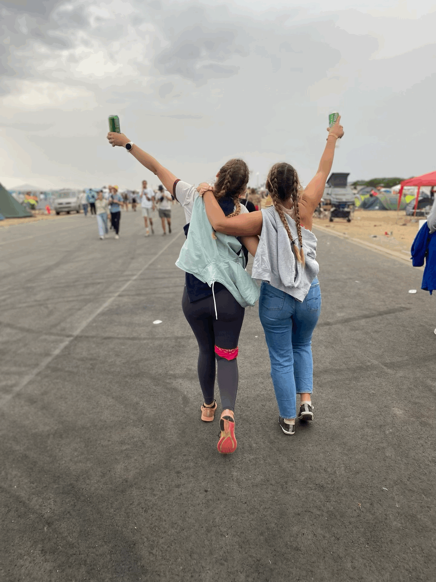 Zwei Frauen stehen mit dem Rücken zum Fotografen. Sie stehen sehr nah und haben den Arm umeinander gelegt. Sie laufen eine Landebahn entlang und strecken jeweils die äußere Hand mit einem Bier nach oben. Ihr Haare sind in zwei Zöpfen geflochten und fallen über ihre Rücken. Um ihren Oberkörper sind Jacken bzw. ein Pulli geschlungen. Im Hintergrund sieht man unscharf weitere Menschen.