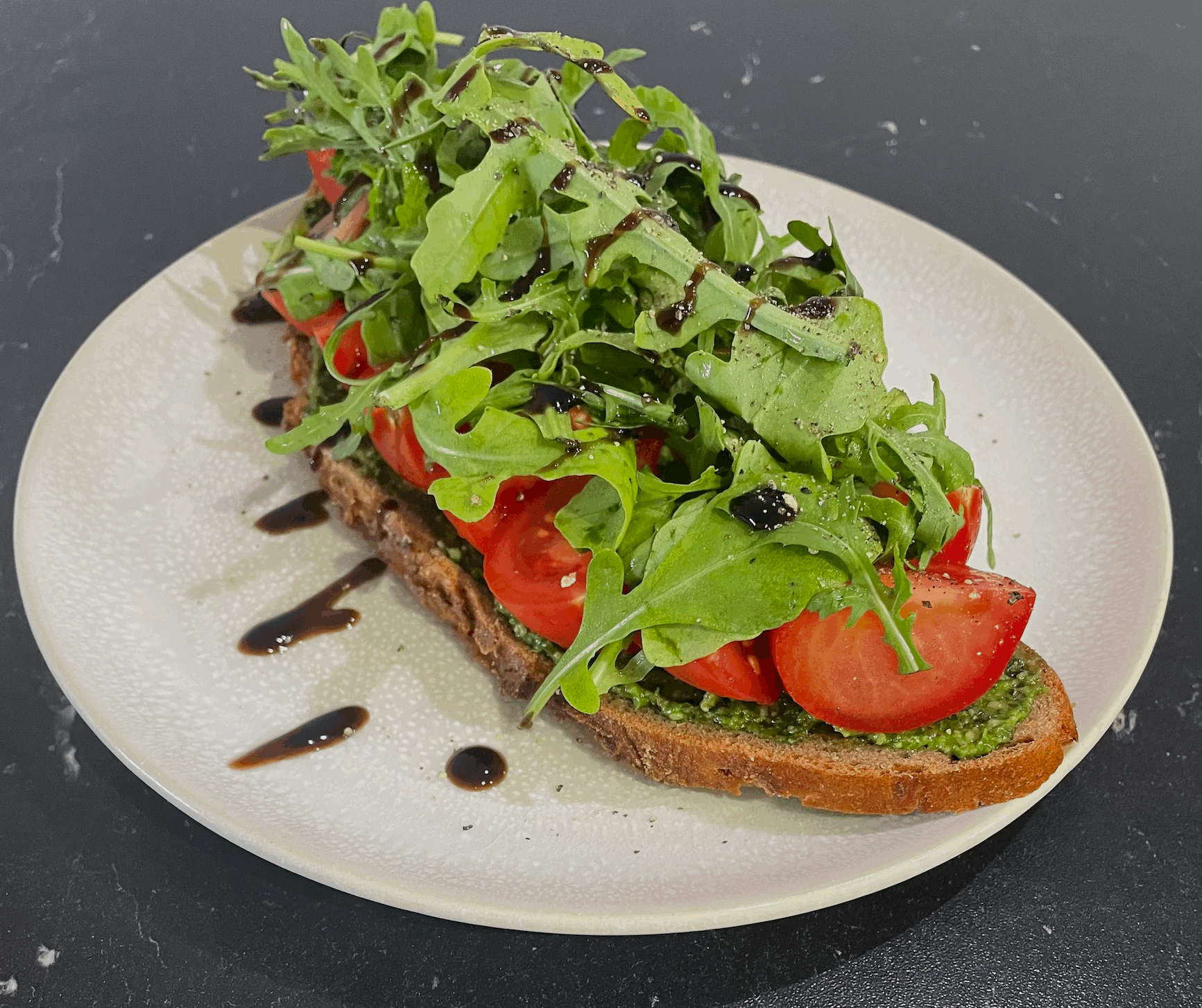 Ein belegtes Brot mit grünem Pesto, Tomaten und Rucola. Verziert mit Creme die Balsamico.