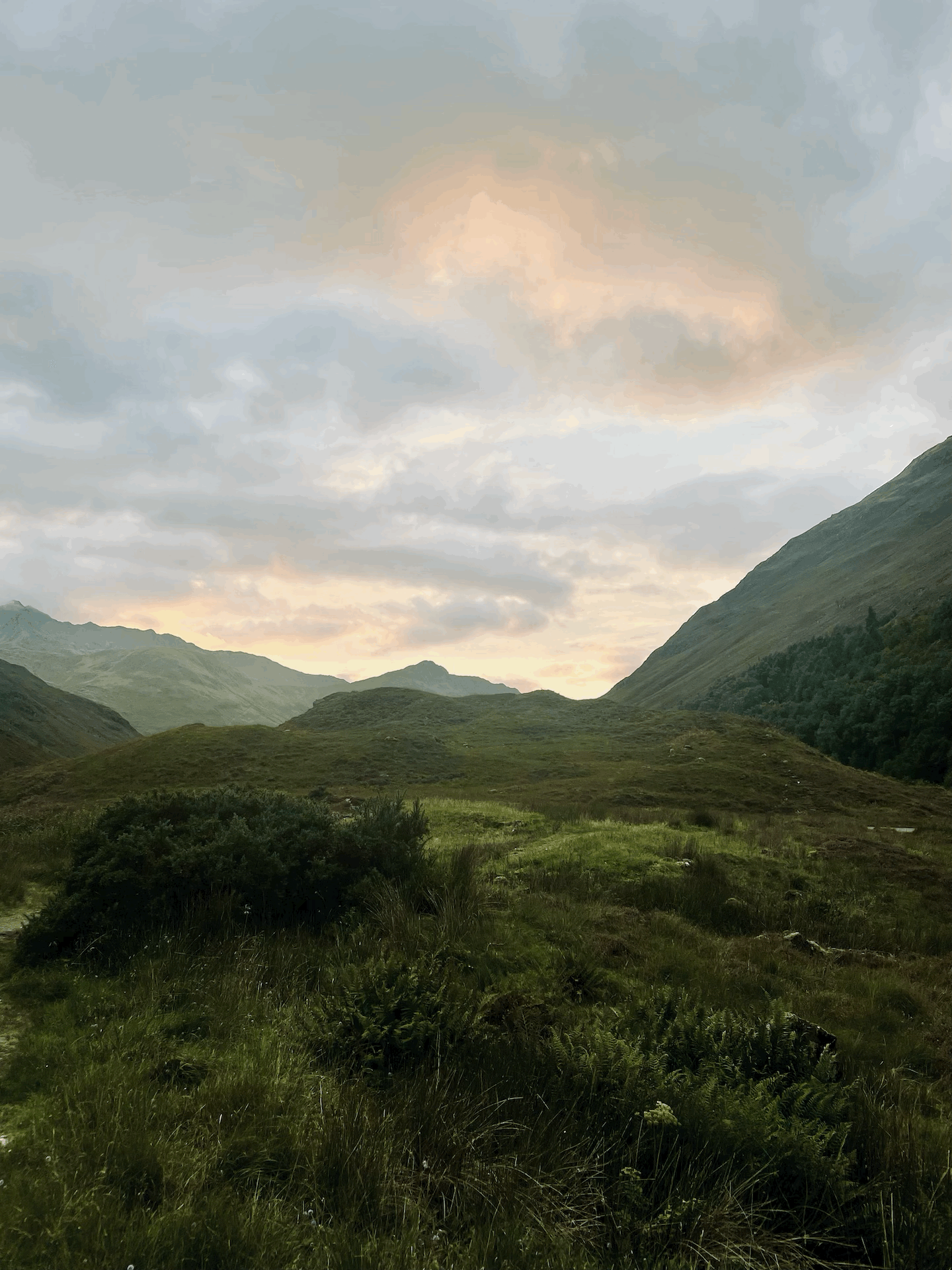 Eine bergige Landschaft. Mit Gras bewachsen. Im Hintergrund sieht man viele Wolken und die Sonne ist gerade dabei unter zu gehen.