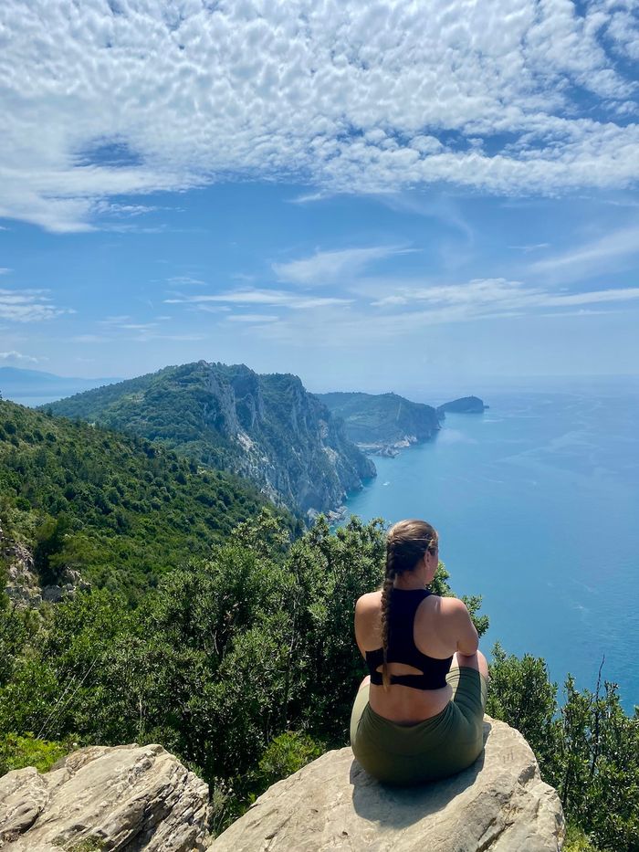 Eine junge Frau in Sport-BH sitzt mit dem Rücken zu uns auf einem Felsen mit Blick auf die Küste der Chinque Terre.