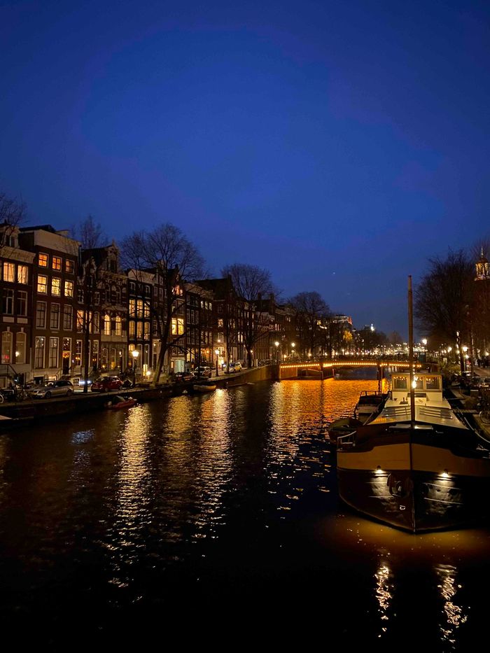 Eine Gracht in Amsterdam. Links gesäumt von beleuchteten Häusern, mittig eine Brücke und rechts ein Boot.